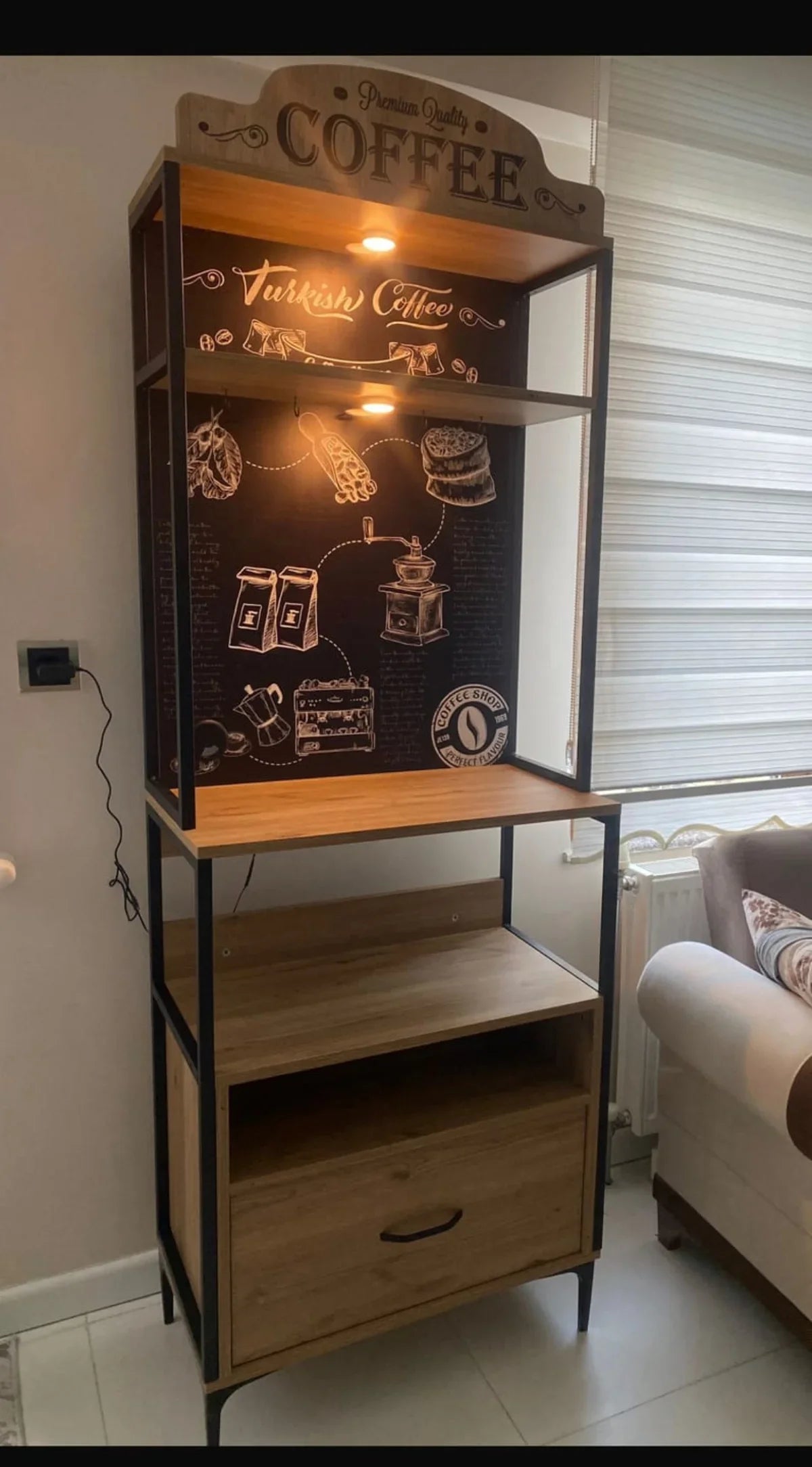 Wooden coffee-themed display cabinet with a light on, placed in a room with a sofa and window blinds.