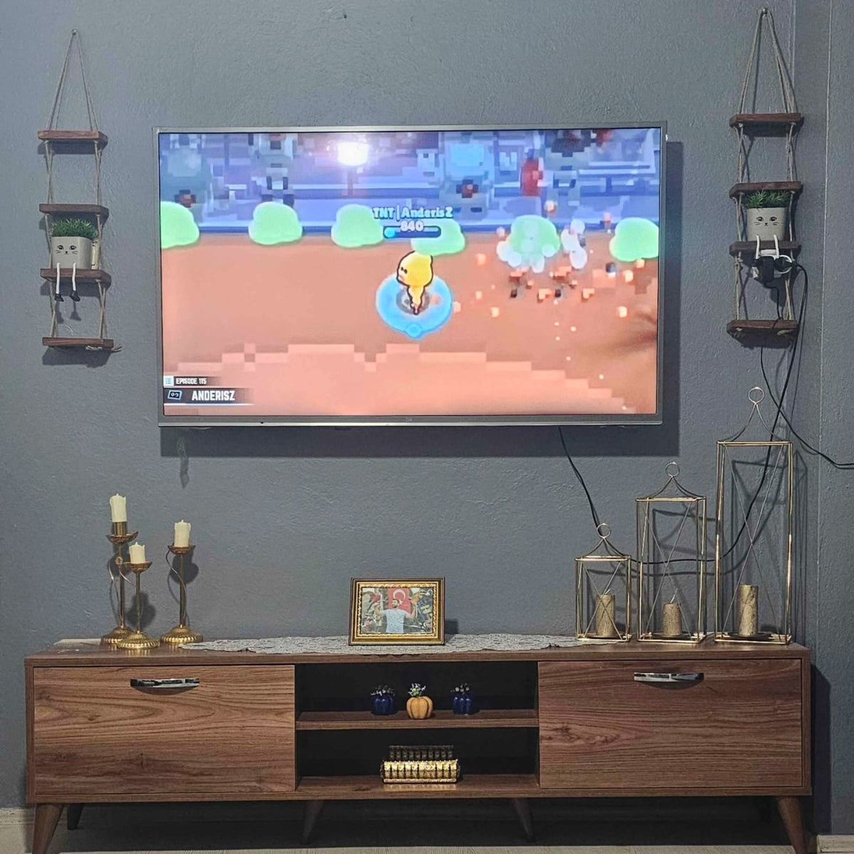 Wooden entertainment console with a TV displaying a game, surrounded by decorative items on a gray wall.