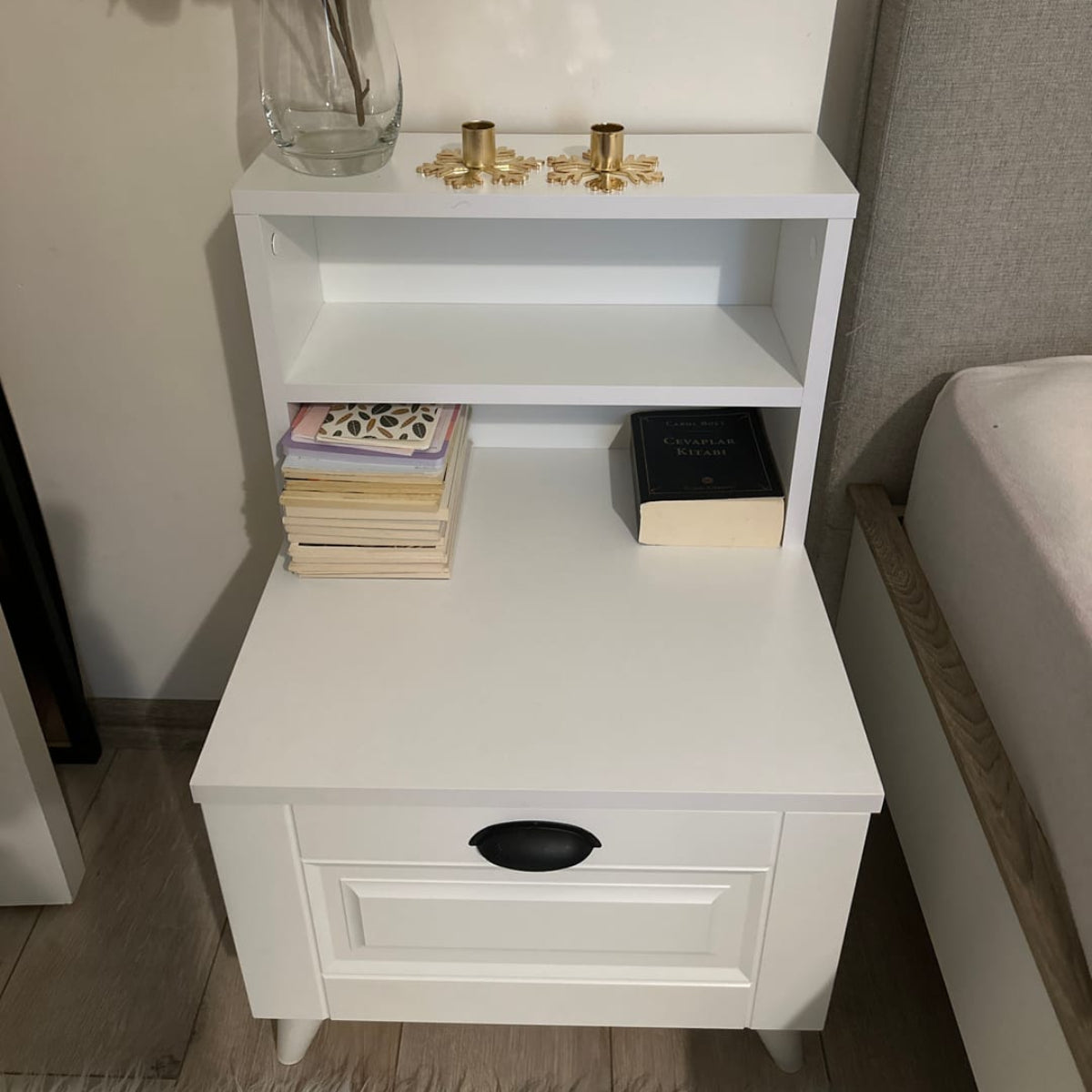 White nightstand with a vase of flowers, books, and decorative items against a neutral wall.