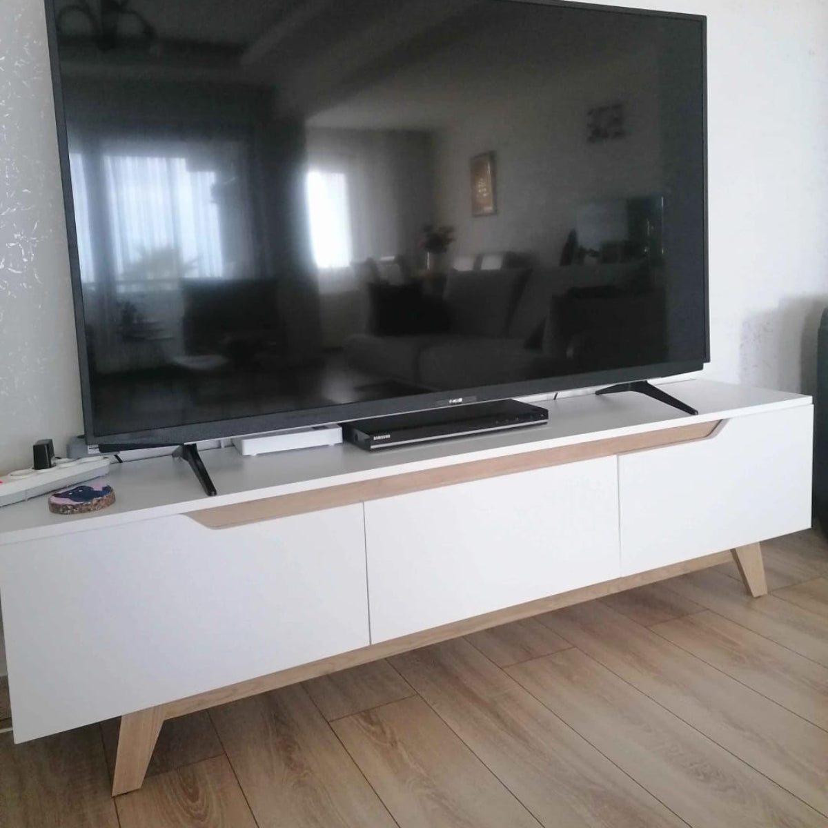 Large flat-screen TV on a white entertainment console with wooden legs in a room.
