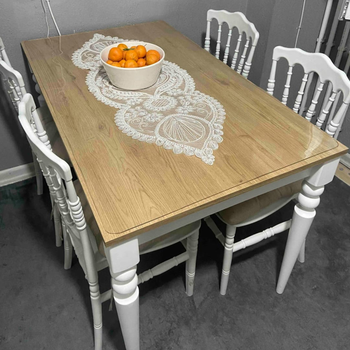 Wooden dining table with white chairs and a bowl of oranges on a gray floor.