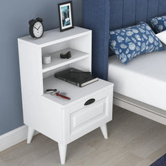 White nightstand with books, clock, and picture frame next to a bed with blue pillows.